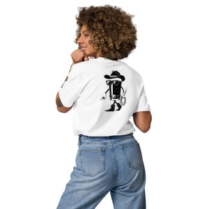 May include: White t-shirt with a black graphic of a cartoon cowboy character on the back. The cowboy is wearing a hat, sunglasses, and boots, holding a lasso. The shirt is made of a soft material.