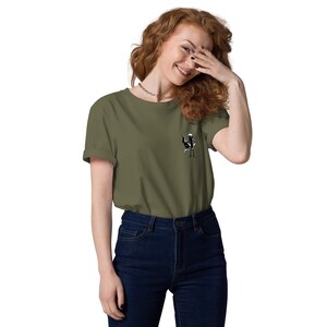 May include: An olive green t-shirt with a small black and white graphic on the chest. The t-shirt has short sleeves and is worn with dark blue jeans. The graphic is a stylized figure.
