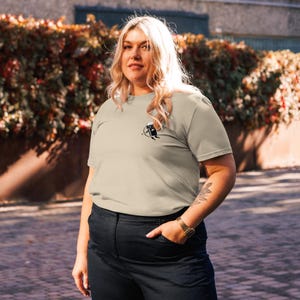 May include: A beige t-shirt with a small black graphic on the chest. The person is wearing black pants and a gold watch. The background features a green bush and a paved walkway.