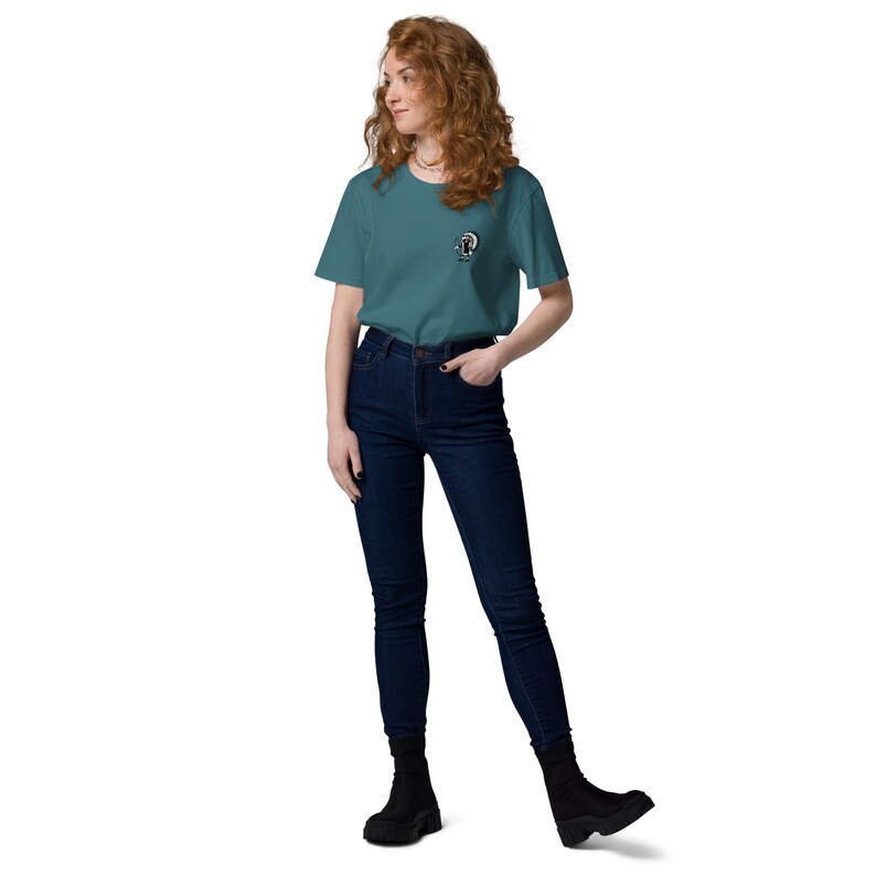 May include: A teal-colored t-shirt with a small graphic on the left chest, paired with dark blue denim jeans. The model is wearing black ankle boots. The t-shirt features a round neck and short sleeves.