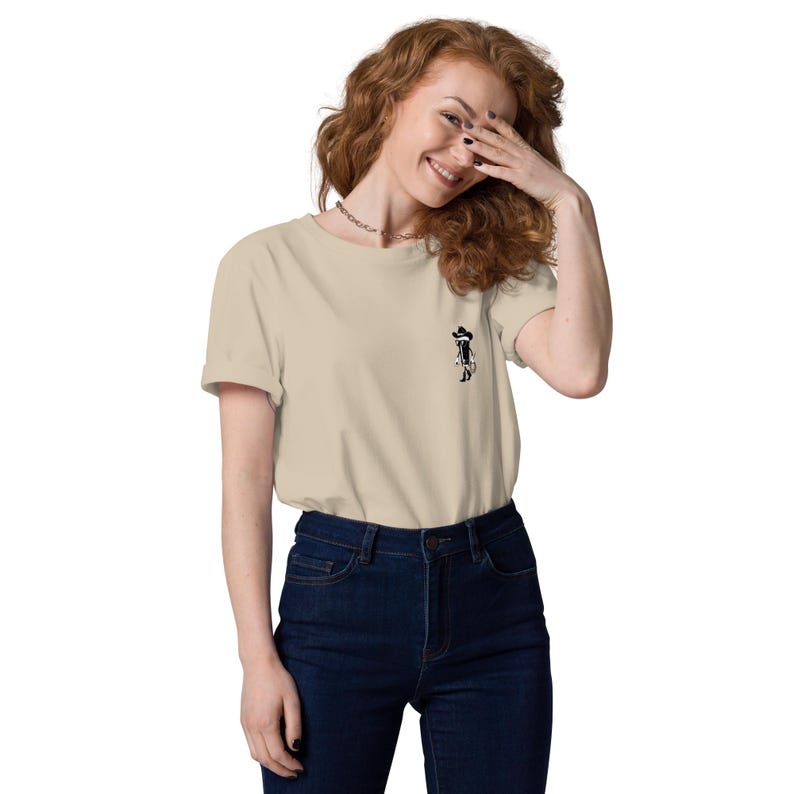 May include: A beige t-shirt with a small black graphic on the front. The graphic appears to be a stylized figure wearing a hat. The shirt is paired with dark blue jeans.