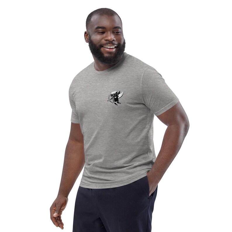 May include: A heather gray t-shirt with a small black and white graphic on the chest. The graphic appears to be a stylized image of a winged object with a bow and arrow. The shirt is short-sleeved and made of a soft material.