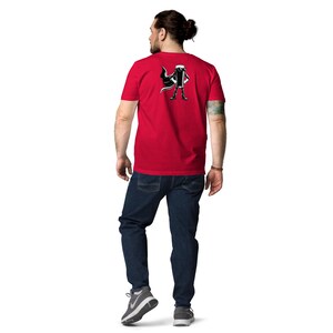 May include: A red t-shirt with a black and white graphic of a character with a cape. The person in the image is wearing blue jeans and grey trainers. The person is facing away from the camera.