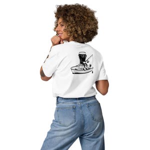 May include: White t-shirt with a black and white graphic of a boat with a person fishing. The person is wearing sunglasses and sitting in a boat with a motor. The t-shirt is made of a soft material.