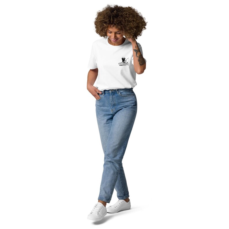 May include: White t-shirt with a small black graphic on the chest. The shirt is paired with light blue jeans and white sneakers. The person is smiling and has curly hair.