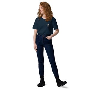 May include: A woman wearing a navy blue t-shirt with a small white graphic on the left chest, paired with dark blue jeans and black boots. The t-shirt features a nautical design. The woman is standing against a white background.