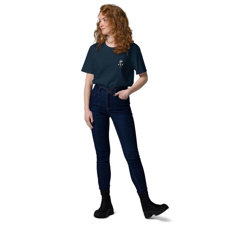May include: A woman wearing a navy blue t-shirt with a small white graphic on the left chest, paired with dark blue jeans and black boots. The t-shirt features a nautical design. The woman is standing against a white background.