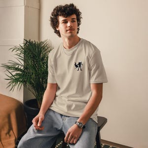 May include: Beige t-shirt with a small black and white graphic on the chest. The shirt has a crew neck and short sleeves. The person is wearing blue jeans and a watch.