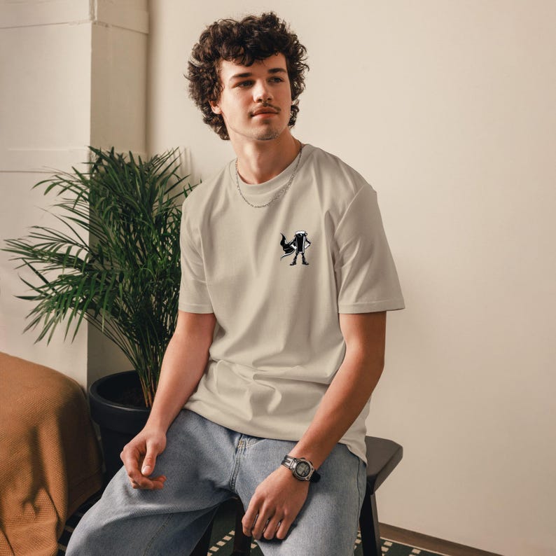 May include: Beige t-shirt with a small black and white graphic on the chest. The shirt has a crew neck and short sleeves. The person is wearing blue jeans and a watch.