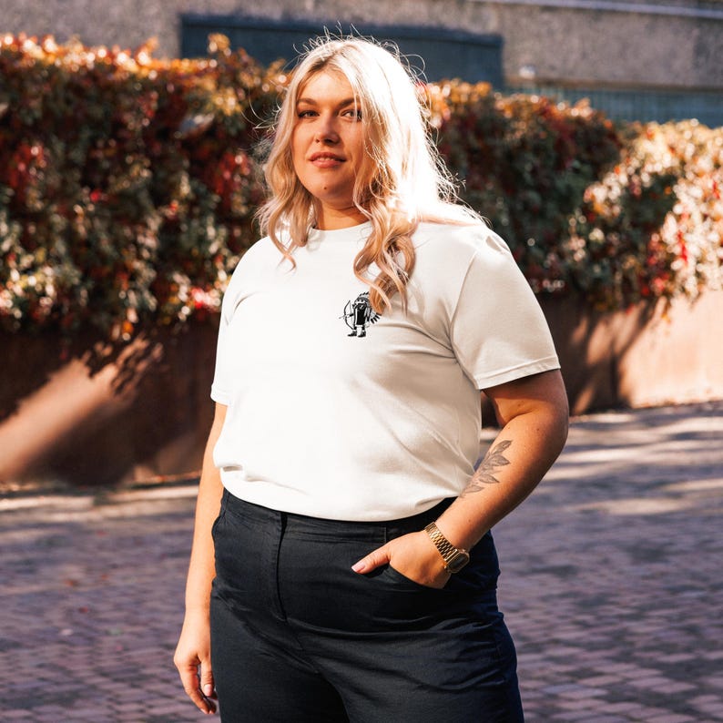 May include: Cream-colored t-shirt with a small black graphic on the chest. The shirt has short sleeves and is paired with dark blue pants. A gold watch is visible on the wrist.