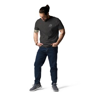 Puede incluir: Camiseta gris oscuro con un peque&ntilde;o gr&aacute;fico blanco en el pecho. La persona lleva vaqueros azul oscuro y zapatillas deportivas grises. La persona est&aacute; de pie con las manos en los bolsillos.