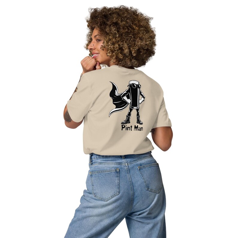 Puede incluir: Camiseta beige con un gr&aacute;fico en blanco y negro de una pinta de cerveza como superh&eacute;roe, con capa y botas. El texto "Pint Man" est&aacute; debajo del gr&aacute;fico. La modelo lleva vaqueros azules.