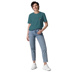 May include: Teal short-sleeved t-shirt with a small graphic on the chest pocket. The shirt is worn with light blue jeans and white trainers. The outfit is casual and suitable for everyday wear.