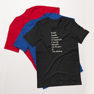 May include: Three t-shirts in red, blue, and dark gray are layered. The dark gray shirt has white text: "Webb, Smails, Noonan, D'Annunzio, Czervik, Spackler, Dr. Beeper, & The Bishop." The shirts are made of soft material.
