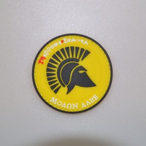 Op de afbeelding: Een ronde gele patch met een zwart Spartaans helm ontwerp. De patch heeft de Griekse zin "ΜΟΛΩΝ ΛΑΒΕ" in het wit en Griekse tekst in rood en wit langs de rand. De patch is ongeveer 7,6 cm in diameter.