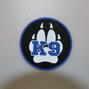 May include: A round patch with a black background, a blue and white paw print, and the blue letters "K9". The patch has a blue border and is likely for law enforcement or military personnel.