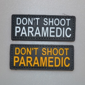 Könnte beinhalten: Zwei rechteckige Aufnäher mit dem Text "DON'T SHOOT PARAMEDIC". Ein Aufnäher hat weißen Text auf schwarzem Hintergrund, der andere gelben Text auf schwarzem Hintergrund. Beide Aufnäher haben einen schwarzen Rand.