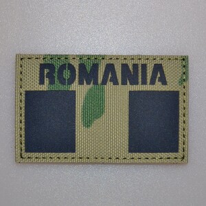 Pode incluir: Um patch retangular com a palavra "ROMANIA" em letras pretas. O patch tem um padrão de camuflagem em tons de verde e bege. Dois quadrados pretos estão abaixo do texto. O patch é contornado com costuras douradas.