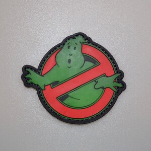 May include: A circular patch featuring the Ghostbusters logo. The design includes a green ghost figure inside a red circle with a diagonal red line, all outlined in black. The patch is likely for clothing or accessories.