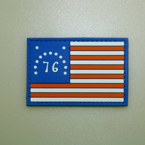 May include: Rectangular patch featuring a stylized American flag design. The flag has a blue field with white stars and the number "76", and red and white stripes. The patch is outlined in blue.