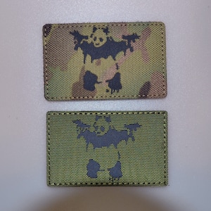May include: Two rectangular patches. The top patch has a camouflage pattern with a black panda-bat hybrid design. The bottom patch is olive green with the same black panda-bat design. Both patches have black stitching.