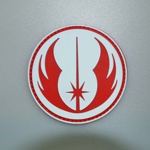 May include: A round, white sticker featuring a red symbol with a central vertical line and a starburst at the bottom. The symbol is framed by red wings and a red border. The sticker is likely related to a science fiction theme.