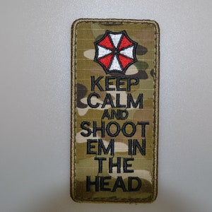 Può includere: Toppa rettangolare con motivo mimetico e logo ombrello rosso, bianco e nero. La toppa presenta il testo nero "KEEP CALM AND SHOOT EM IN THE HEAD".