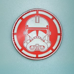 May include: A round, red and white patch featuring a Stormtrooper helmet design. The patch has a white outline and a red border with a stitched edge. The background is a light blue color.