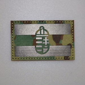 Pode incluir: Remendo retangular com um padrão de camuflagem, uma faixa prateada e um emblema prateado. O emblema é um desenho em forma de escudo com linhas verticais e uma cruz. O remendo é contornado com costura verde escuro.