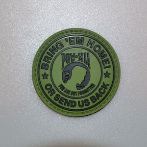 May include: An olive green circular patch with black text. The text around the edge reads "BRING 'EM HOME! OR SEND US BACK". The centre features "POW-MIA" and a silhouette. The bottom text reads "YOU ARE NOT FORGOTTEN".