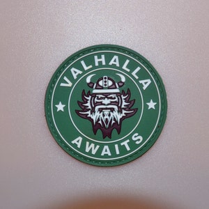 May include: A round, green patch with the words "VALHALLA AWAITS" in white lettering. The patch features a Viking warrior illustration in white and maroon, with a horned helmet and a long beard. Two white stars are on either side of the warrior.