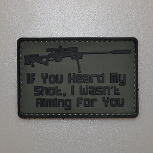 Puede incluir: Parche rectangular verde oliva con un gráfico de rifle de francotirador negro y el texto "IF You Heard My Shot, I Wasn't Aiming For You". El parche tiene un borde negro y probablemente está hecho de tela.