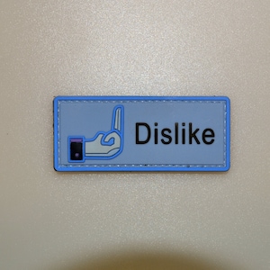 May include: A rectangular patch with a light blue background and a blue border. The patch features a hand gesture with the middle finger raised, outlined in blue, and the word "Dislike" in black text.