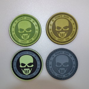 May include: Four circular patches featuring a skull design and the text "DON'T RUN, YOU'LL ONLY DIE TIRED". The patches are in green, tan, and grey. The skull has a mask-like design.