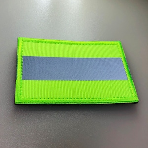 May include: A rectangular patch with a neon green background and a horizontal gray stripe. The patch has a black border and is likely made of woven fabric.