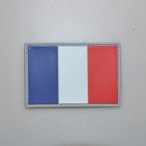 May include: A rectangular patch featuring the French flag design. The patch has three vertical stripes in blue, white, and red, with a gray border. The colors are vibrant and clearly defined, representing the French national flag.