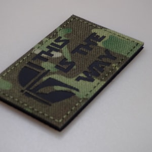 Pode incluir: Remendo retangular com um padrão de camuflagem. O remendo apresenta o texto "THIS IS THE WAY" em letras pretas, juntamente com um gráfico preto de um capacete. As bordas são costuradas com linha bege.