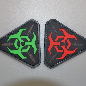 Może przedstawiać: Dwie trójkątne naszywki z czarnym tłem. Jedna naszywka przedstawia zielony symbol zagrożenia biologicznego i brązowe noże, druga czerwony symbol zagrożenia biologicznego i brązowe noże. Obie mają szarą obwódkę.