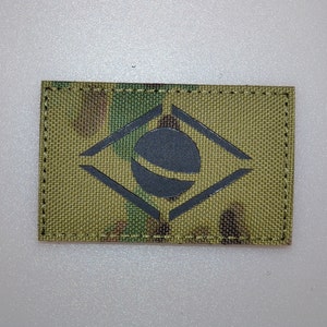 Pode incluir: Um patch retangular com o desenho da bandeira brasileira. O patch tem um fundo de camuflagem e um emblema cinza escuro de um círculo dentro de um losango. As bordas são costuradas com linha escura.