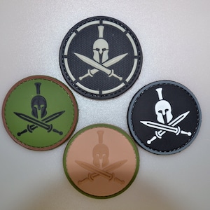 May include: Four round patches featuring a Spartan helmet and crossed swords design. The patches are in various colors: black, green, tan, and black with white accents. The design is a graphic illustration.