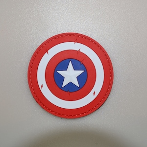 Puede incluir: Parche redondo con el diseño del escudo del Capitán América. El escudo es rojo, blanco y azul, con una estrella blanca sobre un círculo azul en el centro. El borde rojo tiene detalles de rayos blancos.