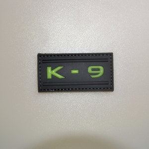 May include: A rectangular black patch with the text "K-9" in bright green. The patch has a stitched border and a textured surface. The letters are raised, giving a 3D effect.