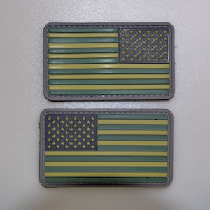 Może przedstawiać: Dwie prostokątne naszywki z wystylizowanym wzorem amerykańskiej flagi. Flaga ma zielone i żółte paski, szare pole gwiazd i szarą obwódkę. Odpowiednie do sprzętu taktycznego.