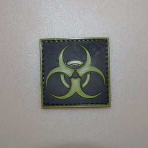 May include: Square patch featuring a biohazard symbol. The symbol is a bright green color, set against a black background. The patch has a green border and is likely made of durable fabric.
