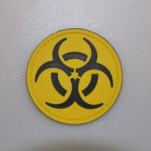 May include: A round, yellow patch featuring a black biohazard symbol. The symbol is centered on the patch, with a black border. The patch is likely made of fabric or a similar material.
