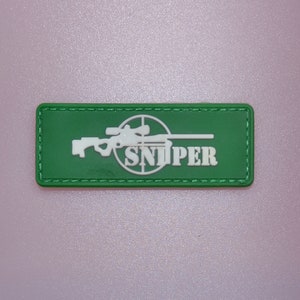 Könnte beinhalten: Ein grünes rechteckiges Abzeichen mit einer weißen Scharfschützengewehr-Grafik und dem Wort "SNIPER" in Weiß. Das Abzeichen hat einen genähten Rand. Der Hintergrund ist hellrosa.