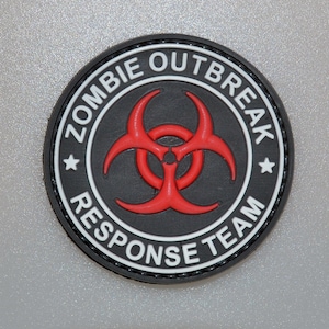 Puede incluir: Un parche redondo negro con texto blanco que dice "ZOMBIE OUTBREAK RESPONSE TEAM". Un símbolo de peligro biológico rojo está en el centro. El parche tiene un borde blanco y dos estrellas blancas.