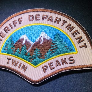 May include: Embroidered patch featuring the words "SHERIFF DEPARTMENT TWIN PEAKS" in brown lettering. The patch has a tan background with a blue sky, brown mountains, and green trees.