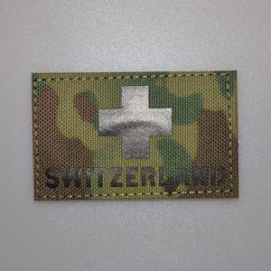 Puede incluir: Un parche rectangular con un patrón de camuflaje en tonos verdes, marrones y beige. Una cruz plateada está centrada, con la palabra "SWITZERLAND" impresa en negro debajo.