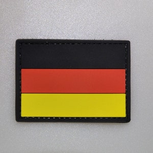 May include: A rectangular patch featuring the German flag design. The flag has three horizontal stripes: black at the top, red in the middle, and yellow at the bottom. The patch is bordered by a black frame.